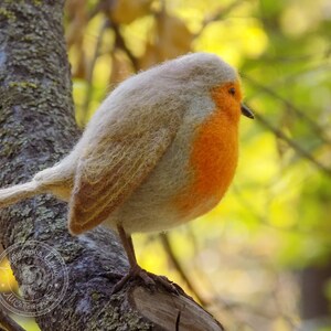 May include: A small, hand-felted robin perched on a branch. The bird is made of wool and has a gray body with an orange breast. The background is blurred and out of focus.