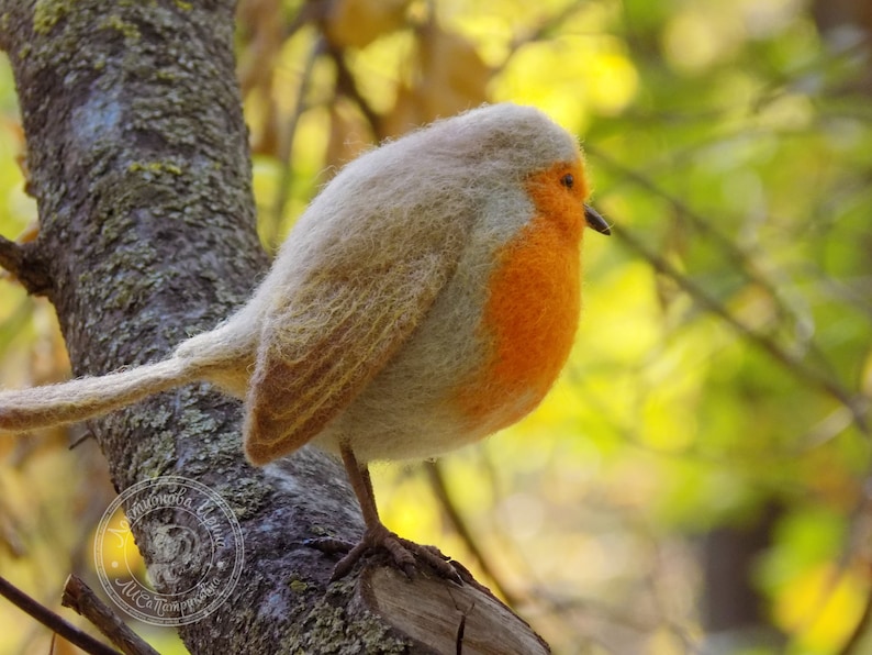 May include: A small, hand-felted robin perched on a branch. The bird is made of wool and has a gray body with an orange breast. The background is blurred and out of focus.