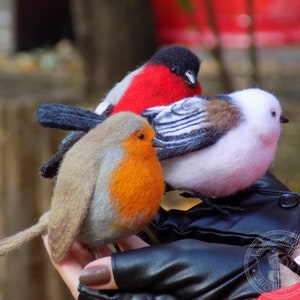 May include: Three felt bird figurines: a red robin, a white and gray bird with black stripes, and a red and black bird. The birds are sitting on a black leather glove.