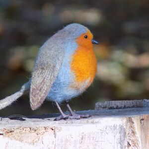 May include: A blue and orange felt robin perched on a weathered tree stump. The robin has a fluffy blue body and a bright orange breast.