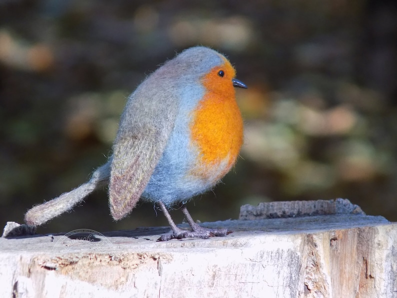 May include: A blue and orange felt robin perched on a weathered tree stump. The robin has a fluffy blue body and a bright orange breast.