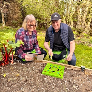 May include: Two people in a garden setting, examining a green planting guide and a green planting tool. A raised garden bed contains soil and rhubarb plants. The people are wearing casual clothing and gardening aprons.