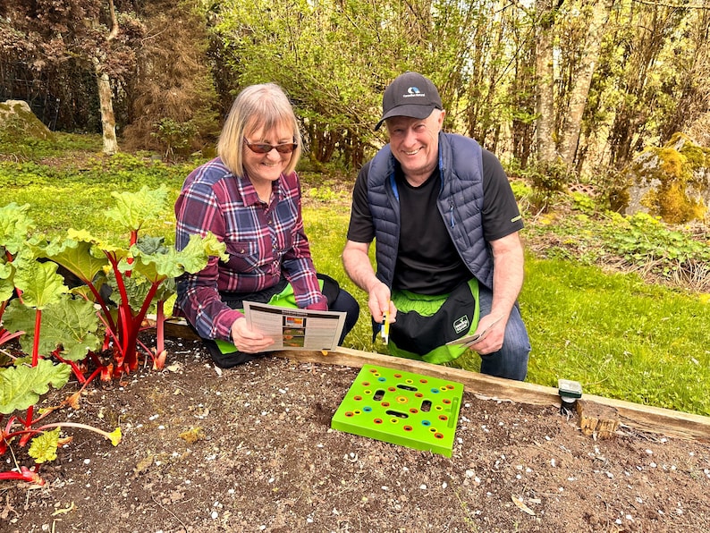 May include: Two people in a garden setting, examining a green planting guide and a green planting tool. A raised garden bed contains soil and rhubarb plants. The people are wearing casual clothing and gardening aprons.