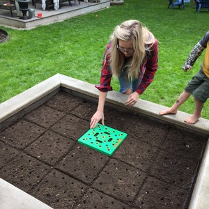 May include: A woman and a child are in a garden bed. The woman is placing a green planting tool on the soil. The garden bed is filled with dark soil and has a concrete border. The child is standing on the border.