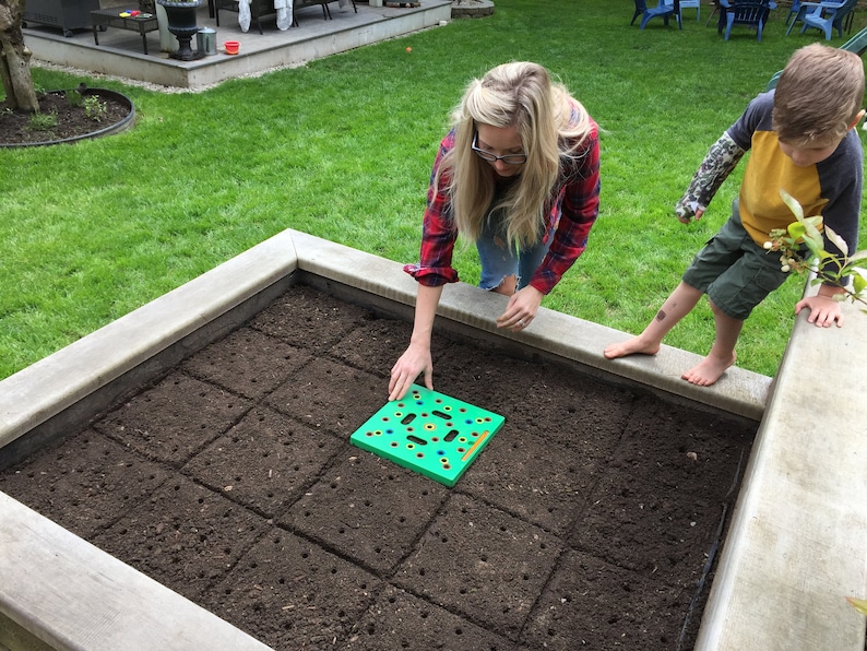 May include: A woman and a child are in a garden bed. The woman is placing a green planting tool on the soil. The garden bed is filled with dark soil and has a concrete border. The child is standing on the border.