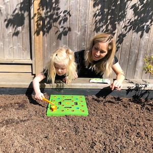May include: A young child and an adult are playing with a green and yellow garden toy in a garden bed. The toy has colorful pegs and a small orange tool. The background includes a wooden fence and a small plant.