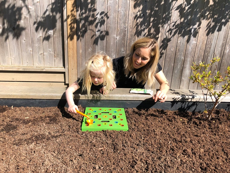 May include: A young child and an adult are playing with a green and yellow garden toy in a garden bed. The toy has colorful pegs and a small orange tool. The background includes a wooden fence and a small plant.