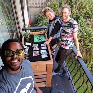 May include: Three people on a balcony with a wooden planter box. The box contains soil, a small plant, and seed packets. One person wears sunglasses. The balcony has a black metal railing and overlooks greenery.