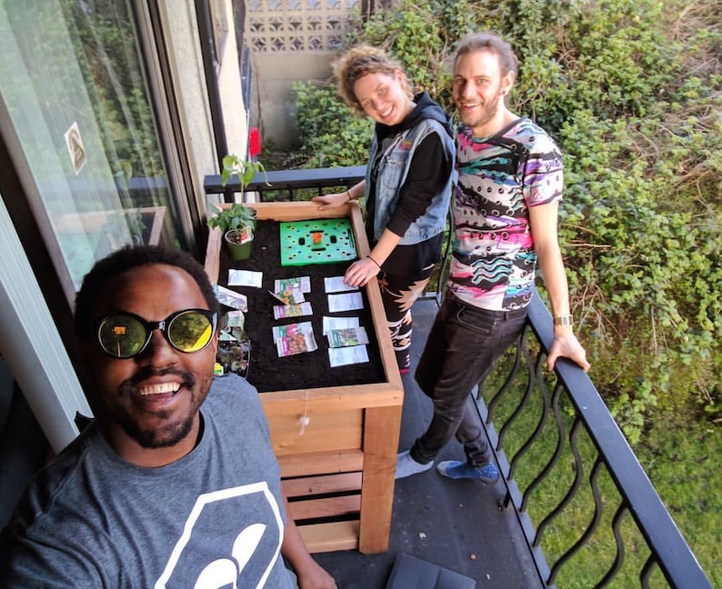 May include: Three people on a balcony with a wooden planter box. The box contains soil, a small plant, and seed packets. One person wears sunglasses. The balcony has a black metal railing and overlooks greenery.