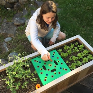 May include: A young person is shown gardening in a wooden planter box. They are using a green planting guide with colored holes to plant seeds. The planter box contains young lettuce plants and other greenery. The person is wearing a white hoodie.