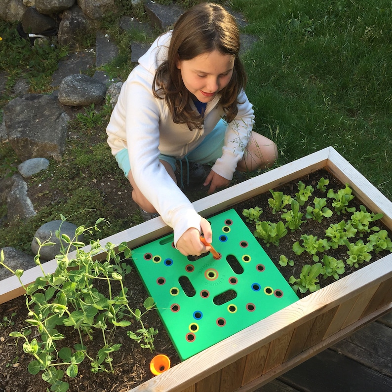 May include: A young person is shown gardening in a wooden planter box. They are using a green planting guide with colored holes to plant seeds. The planter box contains young lettuce plants and other greenery. The person is wearing a white hoodie.
