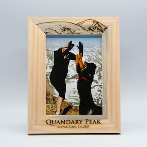 May include: A wooden picture frame with a photo of a person and a dog high-fiving on a mountain peak. The frame has the words "Quandary Peak" and "Elevation: 14,265'" engraved on the bottom.