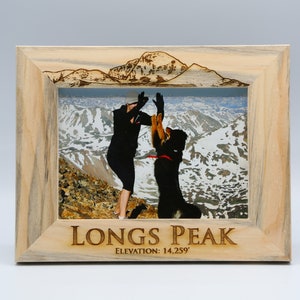 May include: A wooden picture frame with a photo of a person and a dog high-fiving on a mountaintop. The frame is engraved with "Longs Peak" and "Elevation: 14,259'"