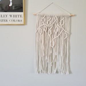 May include: A white macrame wall hanging with a wooden dowel at the top and bottom. The macrame is made with a knot pattern that resembles leaves.