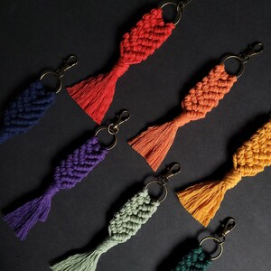 May include: Seven colorful macrame keychains with tassels. The keychains are in shades of red, orange, yellow, green, blue, purple, and teal.