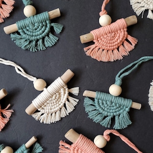 May include: A collection of handmade macrame wall hangings with wooden dowels and beads. The hangings are in various colours, including peach, cream, and teal.