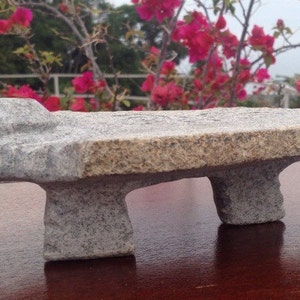 May include: A gray granite stone bench with a textured surface. The bench has a rectangular top supported by two rounded legs. The background features pink flowers and green foliage.