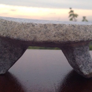 May include: A gray stone mortar and pestle with a rounded top and three legs. The mortar and pestle is sitting on a wooden surface.