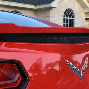 Peut inclure: Gros plan de l'arrière d'une voiture de sport rouge. Comprend une grille en fibre de carbone noire, un aileron et l'emblème argenté Corvette. La finition brillante de la voiture reflète l'environnement.