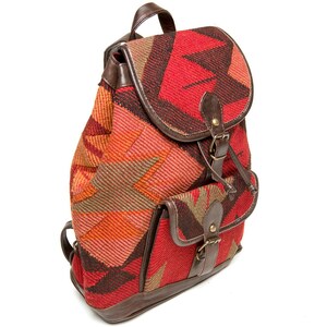 Puede incluir: Mochila colorida con un patrón geométrico en tonos rojos, naranjas y marrones. La mochila tiene un cierre de solapa, dos bolsillos delanteros y detalles en cuero marrón. La bolsa tiene un asa superior y correas ajustables.