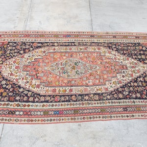 May include: A vintage rug featuring a central medallion in shades of pink, cream, and black. The rug has a geometric pattern with floral motifs and a border of repeating diamond shapes.