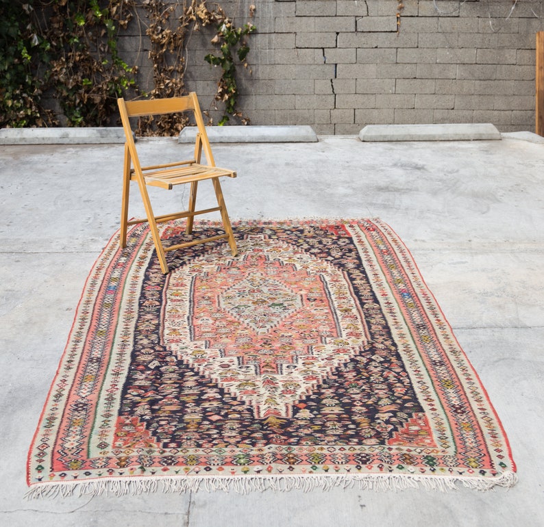 May include: A vintage rug with a dark blue background and a central medallion in shades of pink, cream, and ivory. The rug has a fringe border and intricate geometric patterns.