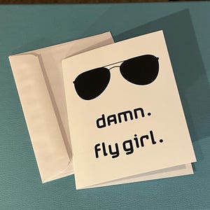May include: A white greeting card with a black sunglasses graphic and the text "damn. fly girl." is next to a white envelope. The card and envelope are on a blue surface.
