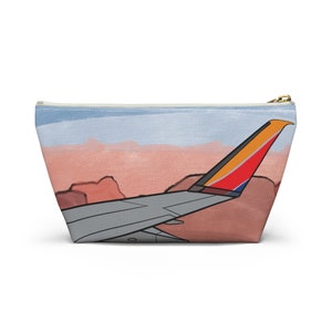 May include: A white zippered pouch featuring a colourful airplane wing design. The wing is painted in shades of red, orange, yellow, and blue. The background is a watercolour-style sky and desert landscape.