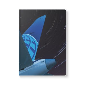 May include: A hardcover notebook featuring a stylized image of an airplane tail fin against a dark night sky with star trails. The tail fin is blue with a white logo. The notebook has a white background.