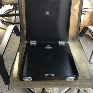 May include: A black metal case with a hinged lid, open to reveal an empty interior. The case is sitting on a beige mesh chair.