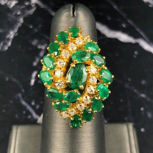 May include: A gold ring featuring a cluster of oval-cut emeralds and small, round diamonds. The emeralds are a vibrant green, and the diamonds sparkle against the gold setting. The ring is displayed on a black finger form.