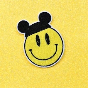 May include: Yellow smiley face with black eyes and a black outline, wearing a black hat with two black mouse ears. The patch is on a yellow glitter background.