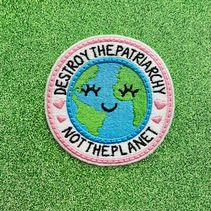May include: Embroidered patch with a smiling Earth design. The patch features a blue and green Earth with closed eyes and a smiling mouth, surrounded by the text "DESTROY THE PATRIARCHY NOT THE PLANET" in a circular pattern. Pink and white accents.