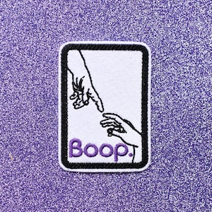 May include: White embroidered patch with a black outline. The patch features two hands reaching towards each other, inspired by the famous "Creation of Adam" painting. The word "Boop" is written in purple below the hands.