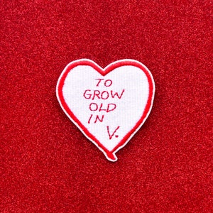 May include: White heart-shaped patch with red stitching and the text "To grow old in V." on a red glitter background.
