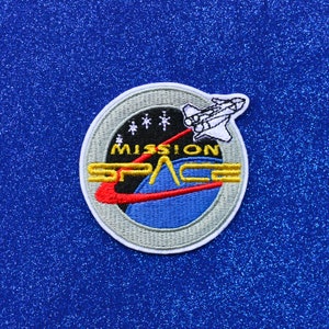 May include: A grey and white embroidered patch with a blue and red design. The patch features a spaceship, stars, and the text "Mission Space".