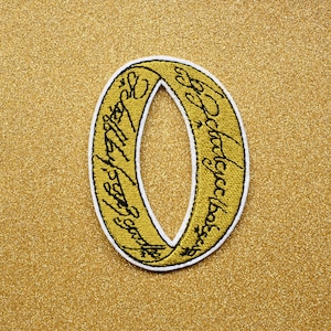 May include: A gold-colored ring with the inscription "One Ring to rule them all, One Ring to find them, One Ring to bring them all and in the darkness bind them" in black lettering.