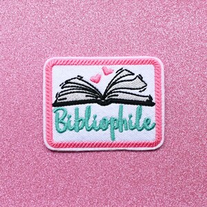 May include: White embroidered patch with a pink border. The patch features an open book with two pink hearts above it and the word "Bibliophile" in teal lettering.