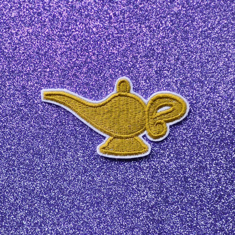 May include: A gold-colored genie lamp patch with white stitching, on a purple glitter background.