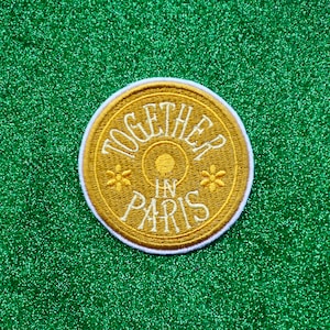 May include: A round, gold embroidered patch with white stitching. The patch says "Together in Paris" with a small circle and two stylized flowers on either side of the text.