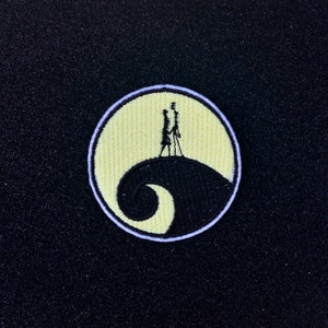 May include: A black and white embroidered patch featuring a silhouette of two figures standing on a hill with a yellow moon behind them. The hill is shaped like a spiral.