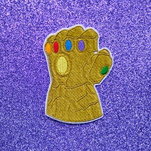 May include: A gold embroidered patch of a hand wearing a gauntlet with six colorful gems. The gauntlet is a popular symbol from the Marvel Cinematic Universe.