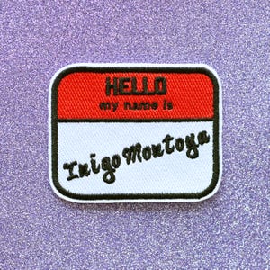 May include: A white name tag with a black border and red top. The tag says "HELLO my name is Inigo Montoya".