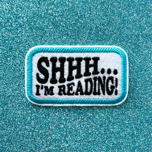 May include: A rectangular embroidered patch with a white background and a bright teal border. The patch features the black embroidered text SHHH... I'M READING! in a bold, sans-serif font, centered within the teal-bordered white rectangular frame.