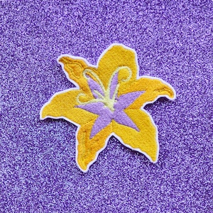 May include: A yellow embroidered lily flower with a purple center, on a purple glitter background.