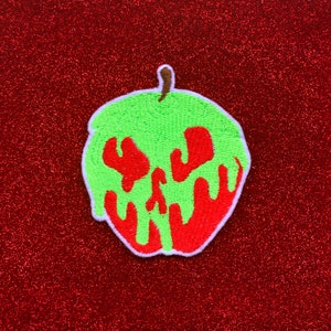 May include: A green apple with a red poison bite mark. The apple is embroidered with a white outline and is on a red glitter background.