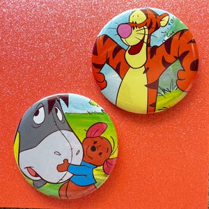 May include: Two circular pin-back buttons featuring cartoon characters. One button displays an orange and black striped tiger with a pink nose. The other button shows a gray donkey being hugged by a small brown creature wearing a blue shirt.