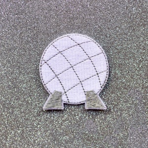 May include: White fabric patch with a stitched design of a sphere with a grid pattern. The sphere is on two small fabric stands.