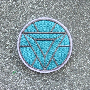 May include: A blue and gray embroidered iron man patch with a white outline. The patch is circular and features a diamond shape in the center.