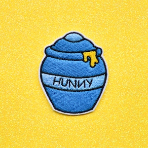 May include: A blue embroidered patch of a honey pot with a yellow drip of honey and the word "Hunny" on the front.
